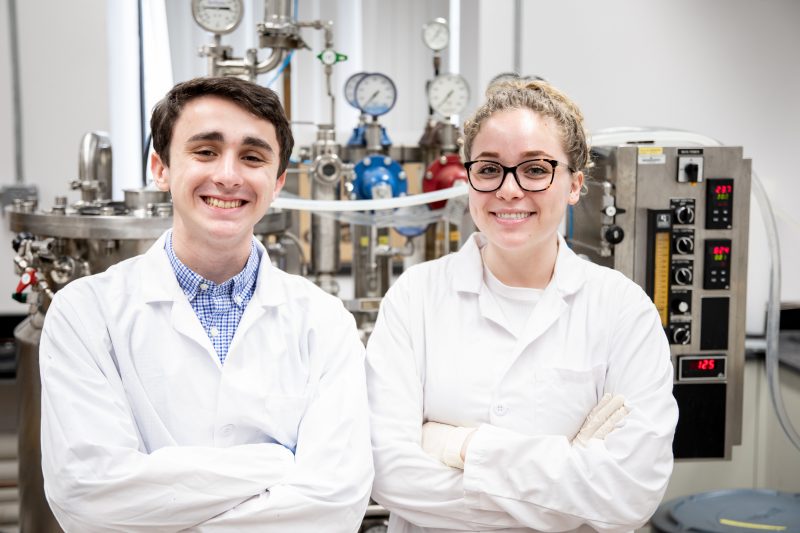Two Virginia Tech Biochemistry students in lab coats smile at the camera with arms crossed