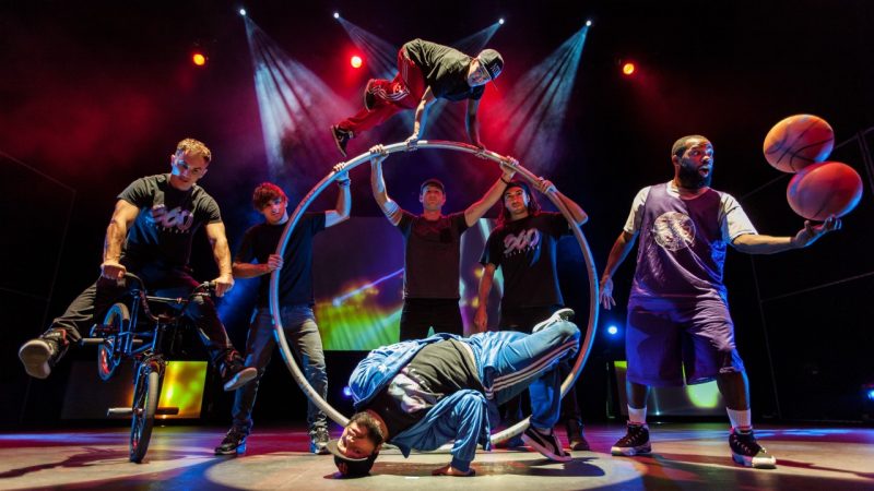 The cast of 360 ALLSTARS gathers on stage. From left, one rides a BMX bike, three hold up a large German wheel while a fourth man balances on top of it, a man hits a breakdance pose, and a man balances one basketball on top of another.