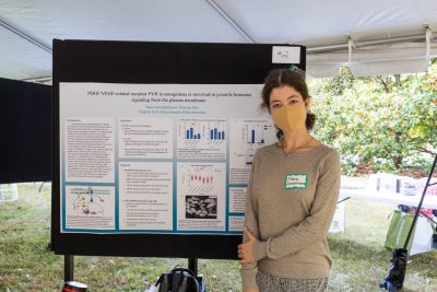 A student stands next to their scientific research poster outside under a tent in a symposium. 