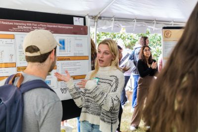 A students gestures towards their scientific research poster and is talking with a student about their research.