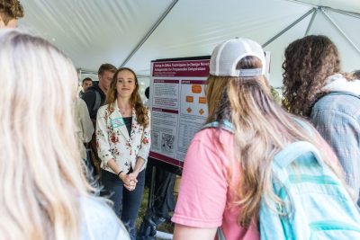 Through a crowd of three students, one student stands next to their scientific research poster and smiles at the crowd.