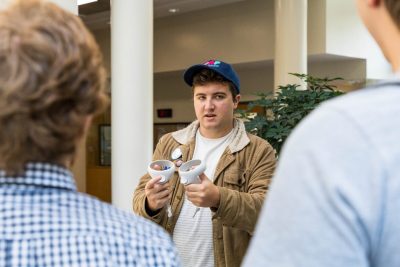 Behind the shoulders of two students, another student in front of them is talking to them and holding up virtual reality hand controllers.