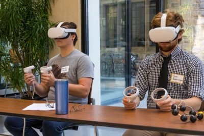 Two students seated inside are wearing virtual reality headsets over their heads and eyes and are holding onto hand controllers.