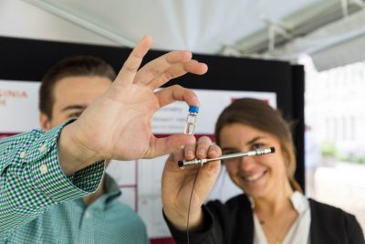 Two students hold up various scientific props and smile towards the camera. The student on the left is holding a small vial with liquid inside while the student on the right is holding a UPLC column.