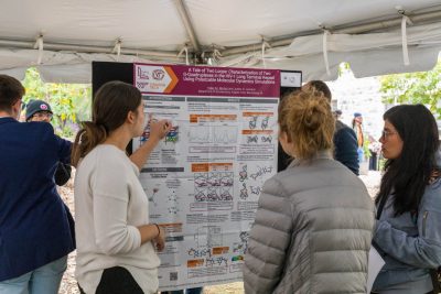 A student presenter points at their scientific research poster while two students listen intently.