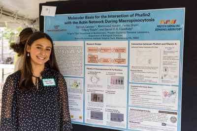 A student stands next to their scientific research poster outside under a tent in a symposium. 