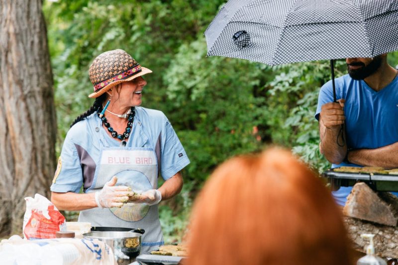 Assistant professor Mae Hey, wearing a hat, braids, and Native-style jewelry, prepares fritters at the Indigenous Friendship Garden.