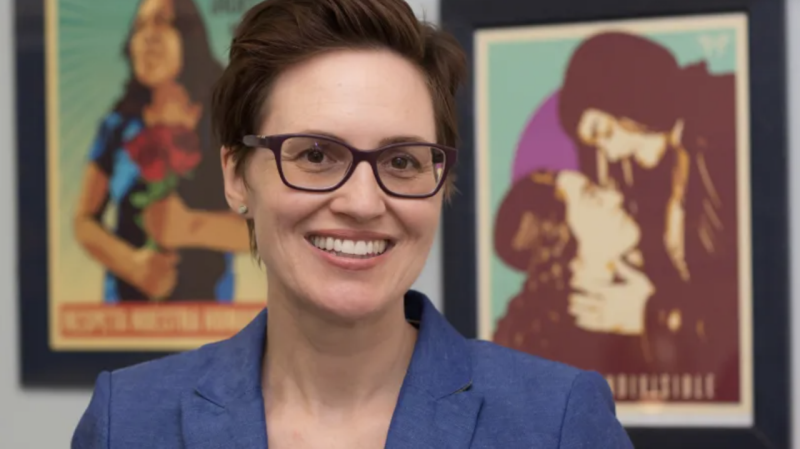 Megan Carney, wearing a blue top, glasses, short hair, stands in front of two colorful posters