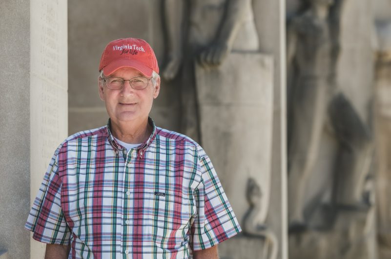 Portrait of Denny Cochrane wearing a plaid button up shirt and a salmon colored hat that reads "Virginia Tech for life." He stands in front of the grey Pylons on a sunny day.